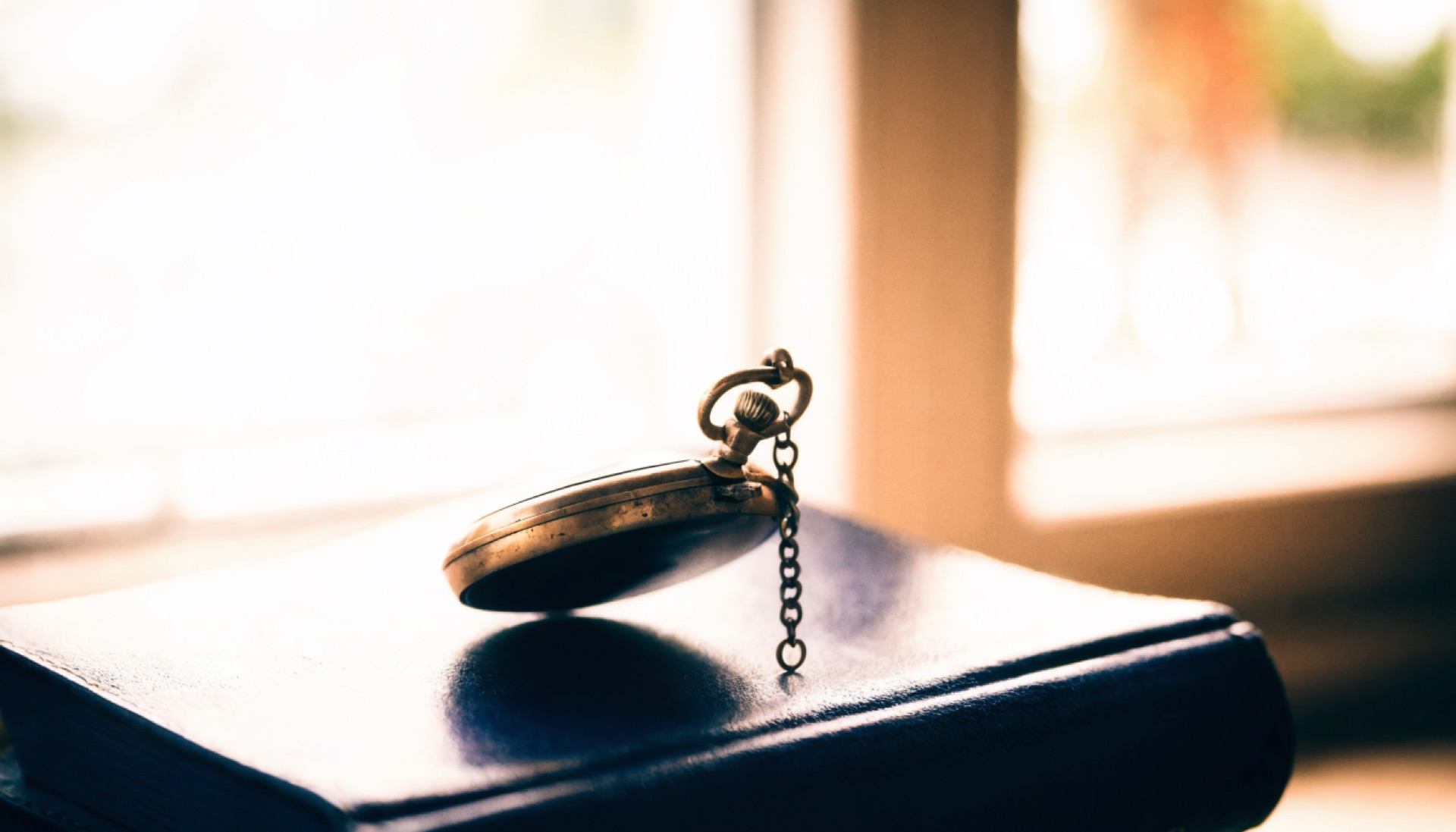 An antique pocket watch resting on a closed leather ledger beside a bright window